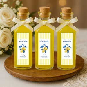 May include: Three small glass bottles filled with yellow liquid, each with a cork stopper and a white ribbon. The bottles have labels that read "Limoncello" and feature a lemon illustration. The bottles are on a wooden tray.