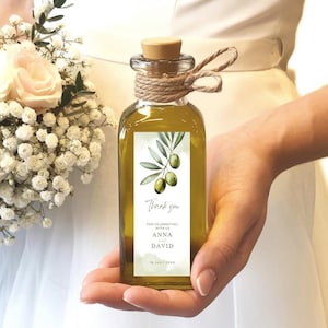 Puede incluir: Una pequeña botella de vidrio transparente llena de aceite de oliva, con tapón de corcho y detalles de cuerda. La botella tiene una etiqueta con ilustraciones de ramas de olivo y el texto "Thank you for celebrating Anna & David". La botella se sostiene frente a un vestido blanco y un ramo.