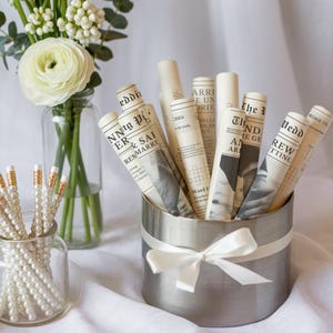 May include: A still-life arrangement featuring pencils with pearl-like beads, rolled paper with newspaper print, and a vase of white flowers. The pencils are in a small glass jar, and the rolled papers are in a silver container with a white ribbon.