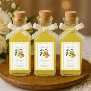 May include: Three small glass bottles filled with yellow liquid, each with a cork stopper and a white ribbon. The bottles have labels with a lemon illustration and the text "Limoncello" and "Thank you". They sit on a wooden tray.