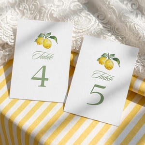 Puede incluir: Dos tarjetas blancas con números de mesa con un diseño de limón en acuarela. Las tarjetas tienen la palabra "Table" en una fuente cursiva y los números 4 y 5 en verde. Están sobre una superficie a rayas amarillas y blancas.