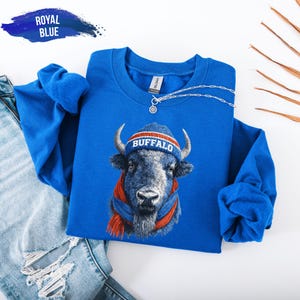 May include: Royal blue sweatshirt featuring a detailed illustration of a buffalo wearing a blue and red knit cap and scarf. The word "BUFFALO" is on the cap. A silver necklace is draped across the sweatshirt. The words "ROYAL BLUE" are in a blue banner in the upper left corner.