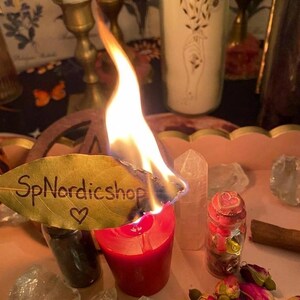 以下が含まれることがあります： 「SpNordicshop」の文字とハートマークが入った葉っぱがついた、燃えている赤いろうそく。ろうそくの周りにはクリスタル、ドライフラワー、小さな瓶が置かれています。背景には、火のついた白いろうそくと花のろうそくがあります。