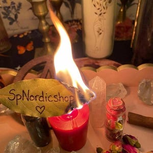 May include: A burning red candle with a leaf that has the text "SpNordicshop" and a heart symbol. The candle is surrounded by crystals, a small jar with a red lid, and dried rose buds. A white candle with floral designs is in the background.