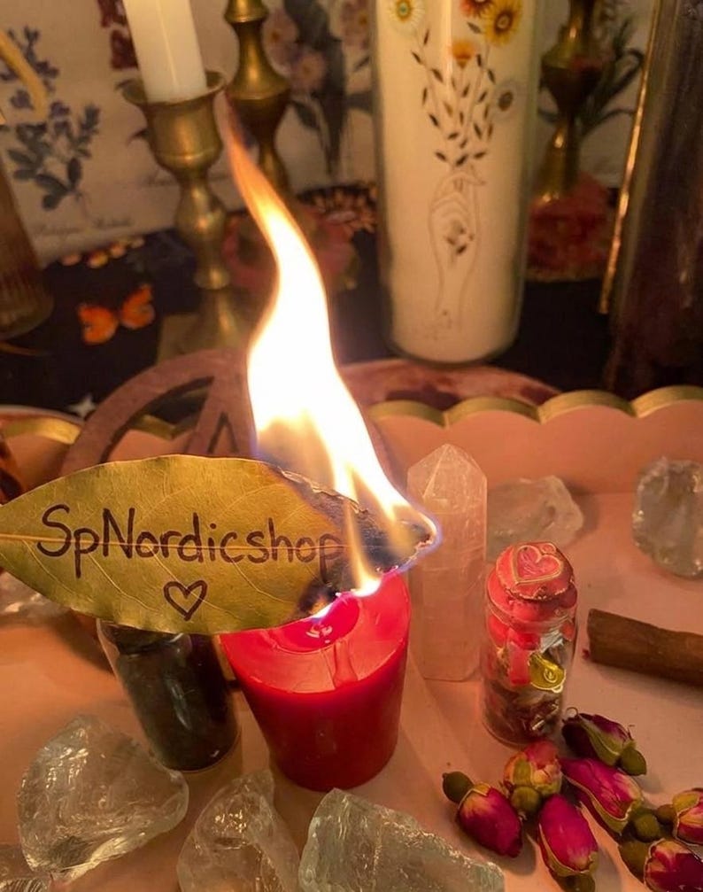 May include: A burning red candle with a leaf that has the text "SpNordicshop" and a heart symbol. The candle is surrounded by crystals, a small jar with a red lid, and dried rose buds. A white candle with floral designs is in the background.