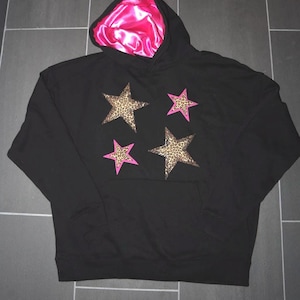 May include: Black hoodie with a pink satin lining on the hood. The hoodie has four star-shaped patches, two are leopard print and two are pink.