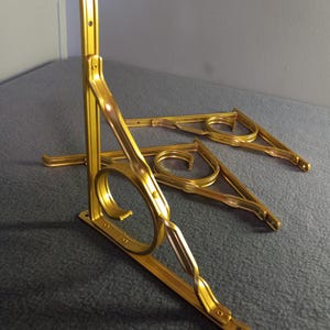 May include: Three gold metal shelf brackets with a decorative scroll design. The brackets are angled and stacked on top of each other.