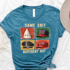 May include: Teal crew neck t-shirt with the text "SAME SHIT DIFFERENT HAT" and images of a KKK hood, a Confederate flag hat, a Nazi hat, and a "Make America Great Again" hat. A tan hat and greenery are in the background.