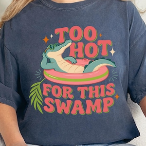 May include: A dark blue t-shirt featuring a graphic of a green alligator wearing sunglasses and lounging in a pink pool float. The text reads "TOO HOT FOR THIS SWAMP" in a retro-style font, accompanied by small orange star accents and a green palm leaf.