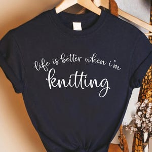 Pu&ograve; includere: T-shirt blu navy con la scritta bianca "life is better when I'm knitting". La maglietta mostra anche la scritta "Custom Infinity Gifts". La maglietta &egrave; appesa a una gruccia di legno.