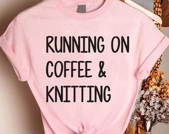 May include: A light pink t-shirt with the text "RUNNING ON COFFEE & KNITTING" in black. The shirt is hanging on a wooden hanger. The words "Custom Infinity Gifts" are printed in a cursive font at the bottom.
