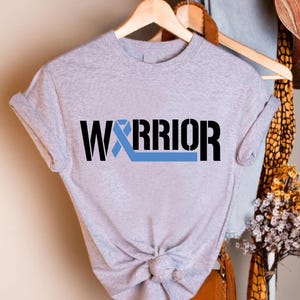 May include: A light gray t-shirt with the word "WARRIOR" in black block letters and a light blue ribbon graphic. The shirt is knotted at the waist and hangs on a wooden hanger.