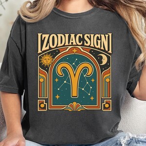 May include: A charcoal gray t-shirt featuring a graphic print with the text ZODIAC SIGN at the top. The design includes a large yellow Aries ram symbol centered within an arched frame, flanked by a sun and moon, set against a dark teal background.