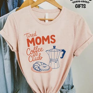 May include: A light pink t-shirt with the words "Tired Moms Coffee Club" in red and blue. The shirt features a graphic of a coffee maker, croissant, and donut. The shirt is hanging on a wooden hanger.