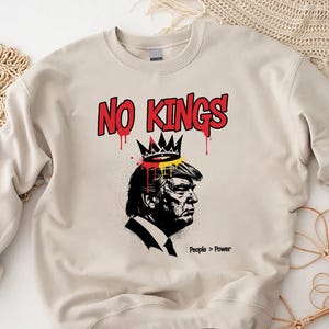 May include: A cream-colored sweatshirt with a graphic design. The design features a black and white profile of a person wearing a crown, with the words "NO KINGS" in red above. The phrase "People > Power" is printed below the profile.