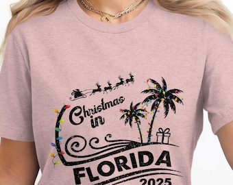May include: Pink t-shirt with a black graphic design. The design features palm trees, a sleigh with reindeer, and the text "Christmas in Florida 2025". Colorful lights and a gift box are also included.