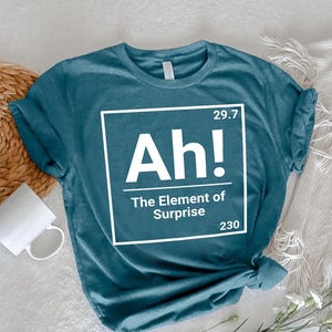 May include: A teal short-sleeved t-shirt featuring a white graphic box designed like a periodic table element. The text inside reads Ah! The Element of Surprise, with the numbers 29.7 and 230 positioned in the top right and bottom right corners.
