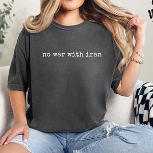 May include: Dark gray t-shirt with the text "no war with iran" in white. The shirt is worn with light blue ripped jeans. The image also includes the text "Custom Infinity Gifts".