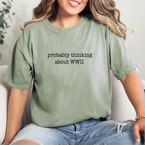 May include: Sage green t-shirt with the text "probably thinking about WWII" in black. The shirt is worn with blue jeans. The words "Custom Infinity Gifts" are printed in a script font.