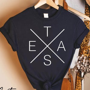 May include: Navy blue t-shirt with the word "TEXAS" in white, arranged in a cross design. The shirt is hanging on a wooden hanger. The words "Custom Infinity Gifts" are printed in black script at the bottom.