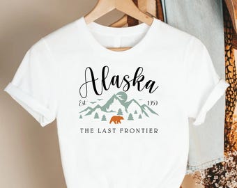 May include: White t-shirt with the word "Alaska" in a script font. Below the word are mountains, trees, and a bear graphic. The text "Est. 1959" and "THE LAST FRONTIER" are also printed on the shirt.