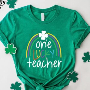 Op de afbeelding: Een groen t-shirt met een regenboog- en klaverontwerp. Het shirt heeft de tekst "one LUCKY teacher" in wit, geel, oranje en blauw. Het shirt is omgeven door klaveruitsparingen.