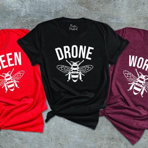 May include: Three t-shirts in red, black, and maroon. Each shirt features a white graphic of a bee and a corresponding word: "QUEEN", "DRONE", and "WORKER". The shirts are short-sleeved and have a casual style.