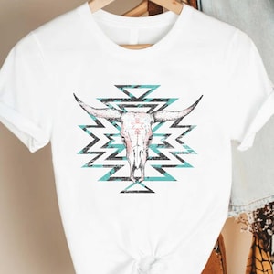 May include: White t-shirt with a turquoise and black geometric design featuring a white bull skull in the center.