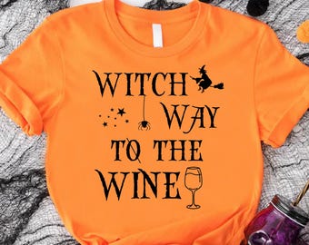May include: Orange t-shirt with the text "Witch Way To The Wine" in black. The design includes a witch on a broom, stars, a spider, and a wine glass. The shirt is knotted at the bottom and is part of a Halloween theme.
