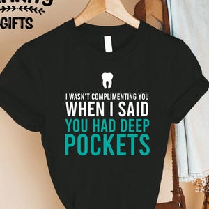 Puede incluir: Camiseta negra con el texto "I WASN'T COMPLIMENTING YOU WHEN I SAID YOU HAD DEEP POCKETS" en blanco y turquesa. Un gr&aacute;fico de diente est&aacute; encima del texto. La camiseta est&aacute; colgada en una percha de madera.