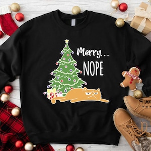 May include: Black sweatshirt with a Christmas tree graphic, a grumpy orange cat, and the text "Merry...NOPE." The tree is decorated with ornaments and lights. Presents are at the base of the tree. A humorous holiday design.