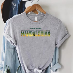 Puede incluir: Una camiseta gris con el texto "STAR WARS THE MANDALORIAN" en verde, amarillo y naranja. La camiseta est&aacute; colgada en una percha de madera, con otras prendas en el fondo.