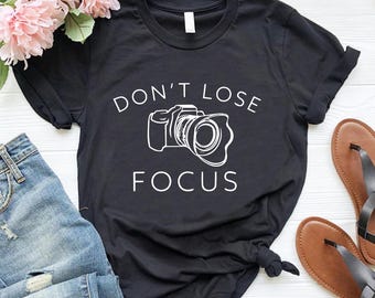 May include: Black t-shirt with the text "DON'T LOSE FOCUS" and a camera graphic in white. The shirt is paired with blue jeans, brown sandals, and pink flowers. The shirt is knotted at the waist.