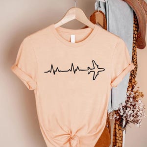 Puede incluir: Camiseta color melocot&oacute;n con un gr&aacute;fico negro de una l&iacute;nea de latidos que se transforma en la silueta de un avi&oacute;n. La camiseta de manga corta est&aacute; colgada en una percha de madera.