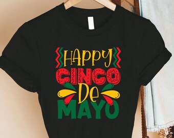 May include: Black t-shirt with a colorful "Happy Cinco De Mayo" graphic. The design features red, green, and yellow text and decorative elements. The shirt is hanging on a wooden hanger.