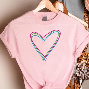 May include: A light pink t-shirt with a multi-colored heart design. The heart is outlined in red, green, and blue. The shirt is hanging on a wooden hanger. The sleeves are cuffed.