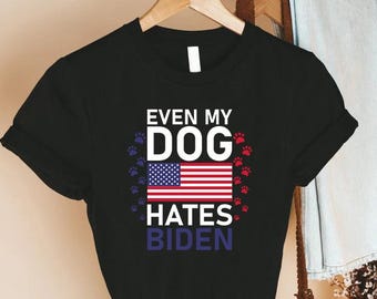 May include: Black t-shirt with the text "EVEN MY DOG HATES BIDEN". The design includes an American flag and paw prints in red and blue. The shirt is hanging on a wooden hanger.