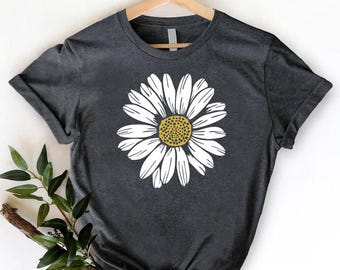 May include: Dark gray t-shirt featuring a large white daisy with a yellow center. The t-shirt is displayed on a wooden hanger against a white background. A sprig of greenery is placed on the lower left side of the shirt.