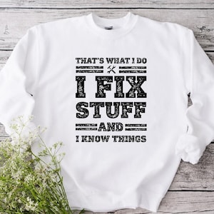 May include: White sweatshirt with the text "THAT'S WHAT I DO I FIX STUFF AND I KNOW THINGS" in black block letters. The design includes a wrench and a hammer graphic. The sweatshirt is displayed on a light wood-grain surface.
