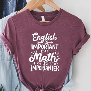 May include: A maroon t-shirt with the text "English is important but Math is more importanter" in white. The shirt is hanging on a wooden hanger with other clothing items in the background.