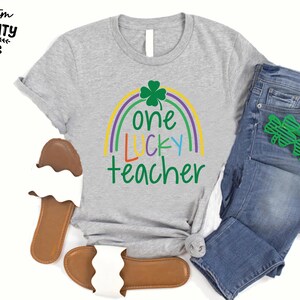 Op de afbeelding: Een grijs t-shirt met de tekst "one LUCKY teacher" in groen, geel, blauw en paars, met een regenboog en klaverontwerp. Het shirt wordt gecombineerd met een blauwe jeans, groene klavervormige brillen en bruine en witte sandalen.