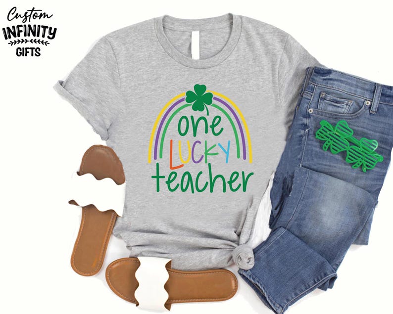 Op de afbeelding: Een grijs t-shirt met de tekst "one LUCKY teacher" in groen, geel, blauw en paars, met een regenboog en klaverontwerp. Het shirt wordt gecombineerd met een blauwe jeans, groene klavervormige brillen en bruine en witte sandalen.