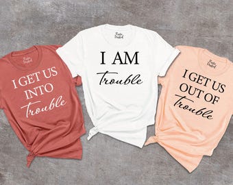 May include: Three short-sleeved t-shirts in coral, white, and peach. The coral shirt reads "I GET US INTO Trouble", the white shirt says "I AM Trouble", and the peach shirt says "I GET US OUT OF Trouble". All shirts have black text.
