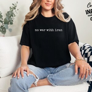May include: Black t-shirt with the text "no war with iran" in white. The shirt is worn by a person sitting on a white couch. The words "Custom Infinity Gifts" are printed on the upper right corner.