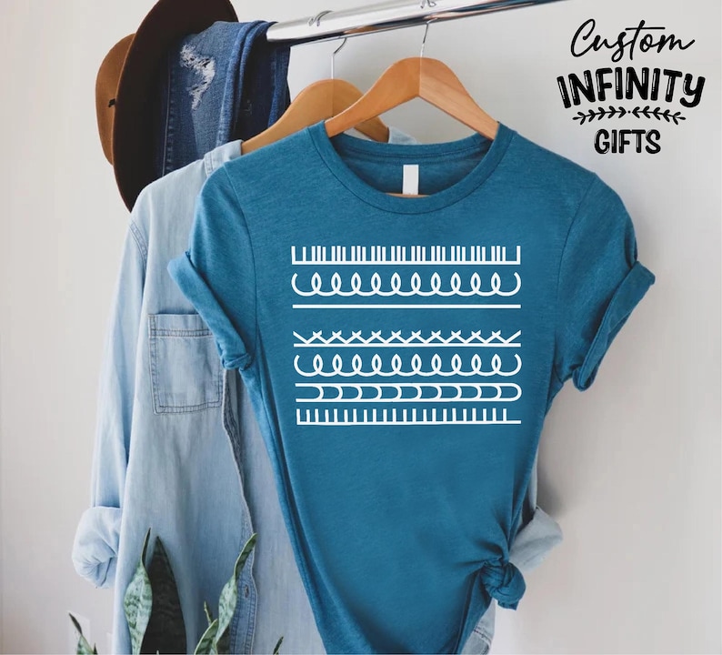 May include: Teal t-shirt with white graphic designs, hanging on a wooden hanger. The shirt features various white line patterns. The text "Custom Infinity Gifts" is in the upper right corner. A denim shirt and a brown hat are also visible.