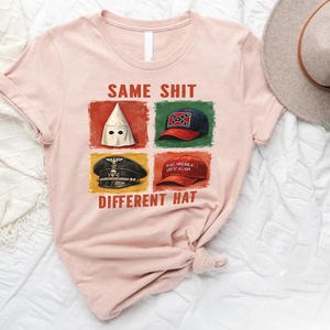 May include: A light pink t-shirt with the text "SAME SHIT DIFFERENT HAT" and images of a white hood, a Confederate flag hat, a Nazi hat, and a "Make America Great Again" hat.