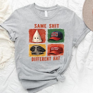 May include: A light gray t-shirt with the text "SAME SHIT DIFFERENT HAT" and images of a KKK hood, a Confederate flag hat, a Nazi hat, and a "Make America Great Again" hat. The shirt is on a white surface.