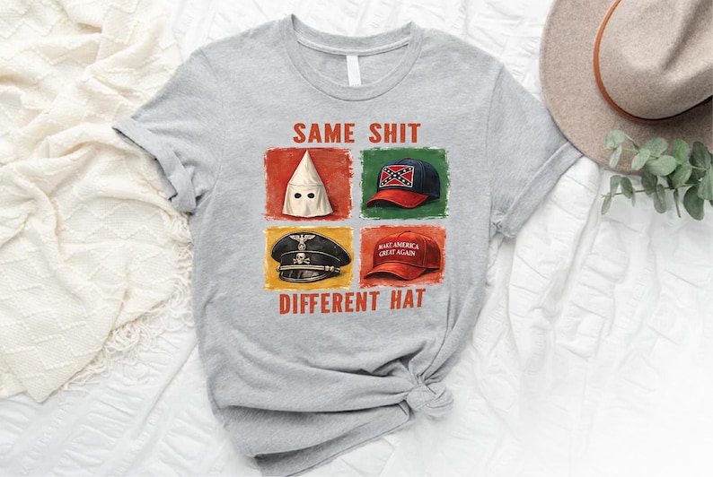 May include: A light gray t-shirt with the text "SAME SHIT DIFFERENT HAT" and images of a KKK hood, a Confederate flag hat, a Nazi hat, and a "Make America Great Again" hat. The shirt is on a white surface.