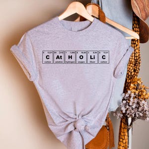 Peut inclure: Un t-shirt gris clair avec le mot "Catholic" &eacute;pel&eacute; avec les &eacute;l&eacute;ments du tableau p&eacute;riodique. Le t-shirt est nou&eacute; &agrave; la taille et est suspendu &agrave; un cintre en bois.