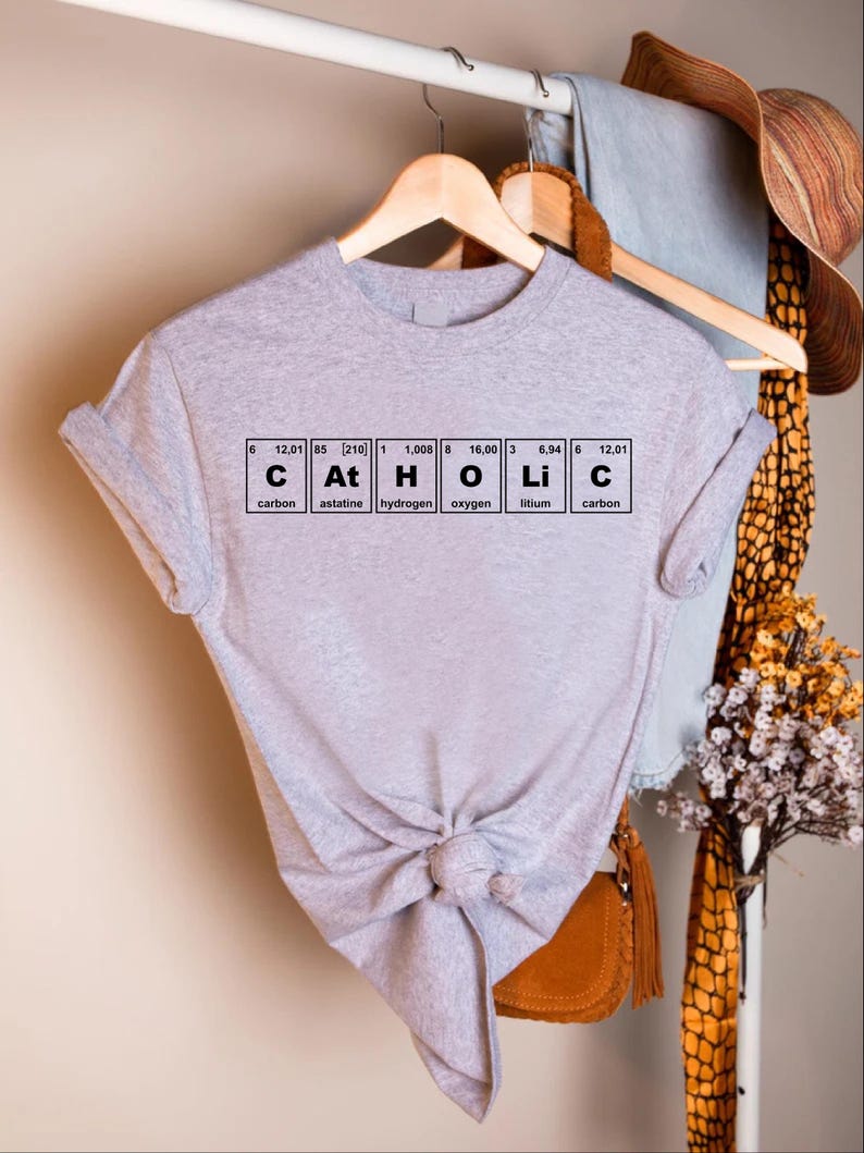 Peut inclure: Un t-shirt gris clair avec le mot "Catholic" &eacute;pel&eacute; avec les &eacute;l&eacute;ments du tableau p&eacute;riodique. Le t-shirt est nou&eacute; &agrave; la taille et est suspendu &agrave; un cintre en bois.